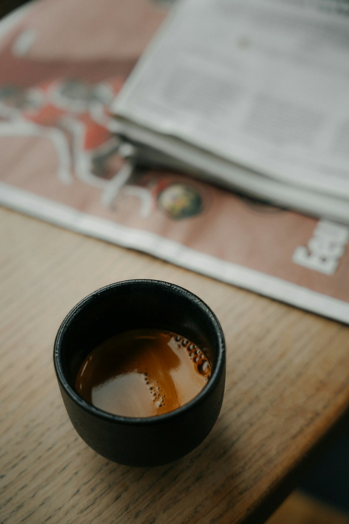 An espresso cup on a wooden table beside a folded newspaper