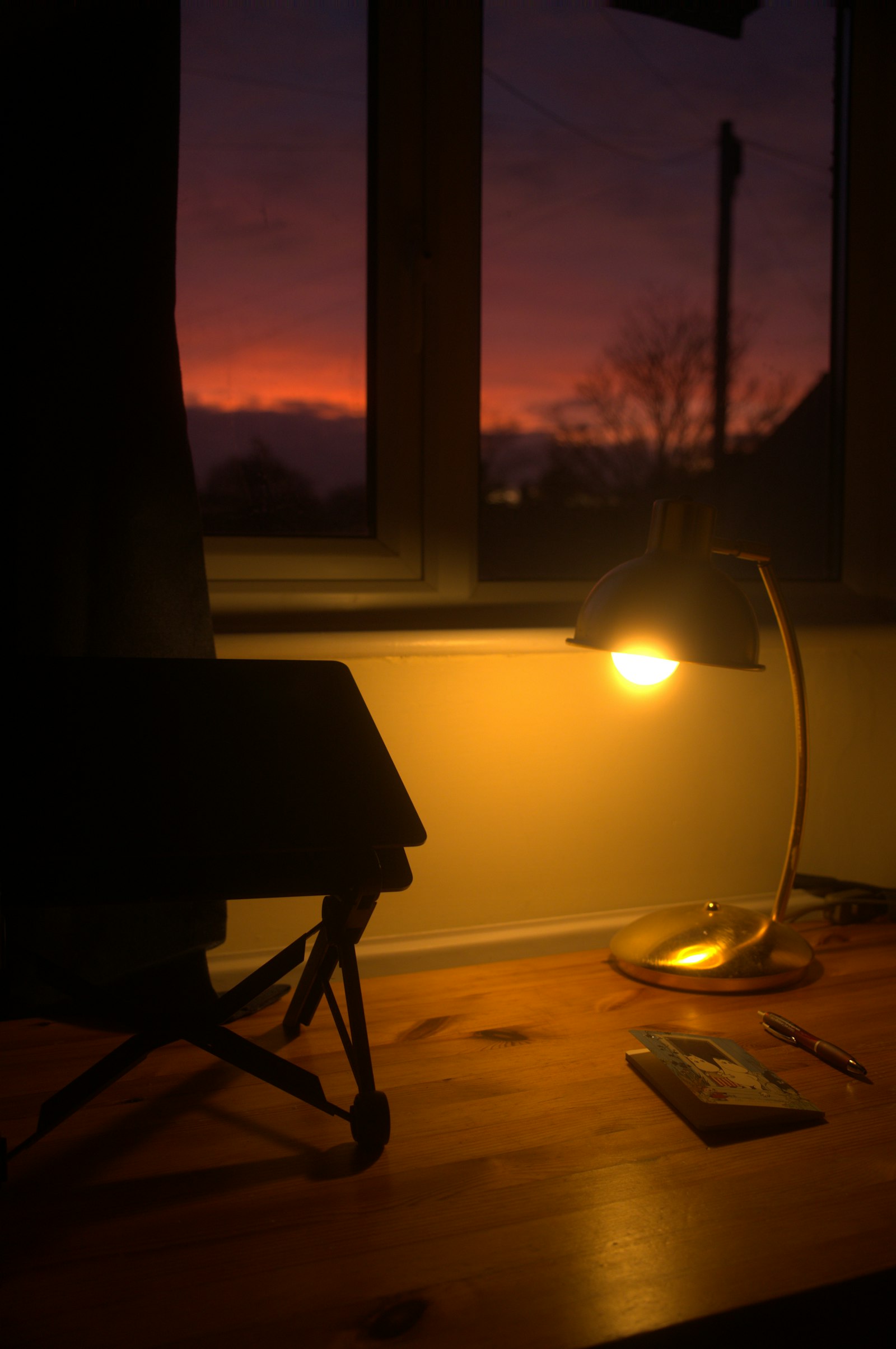A brass desk lamp glowing over a wooden desk at dusk, winter sunset through the window